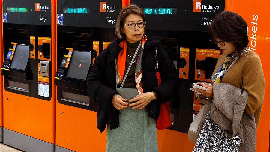 En la estación, dos mujeres se enfrentan a máquinas expendedoras de billetes. Una revisa su bolso y la otra, un billete, reflejando la afectación del servicio.