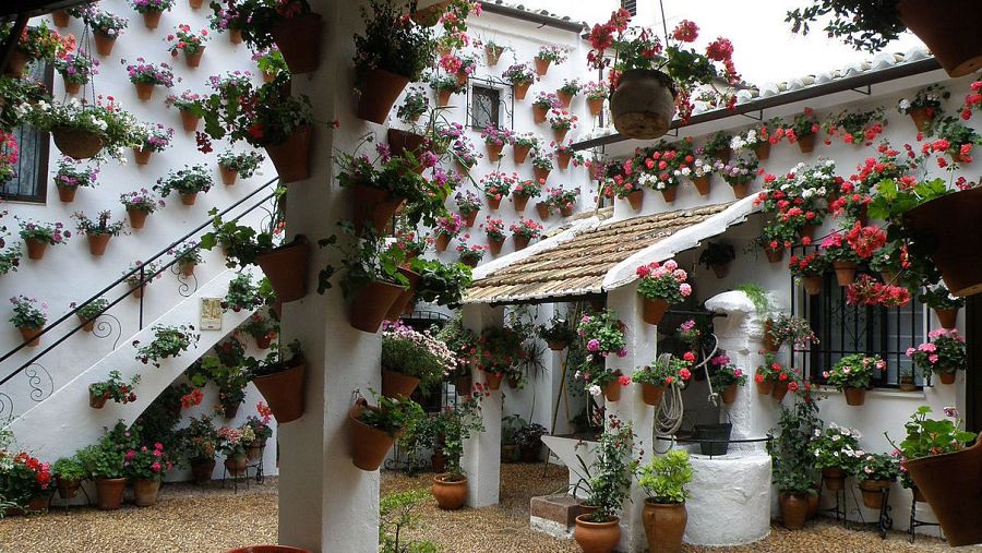 Un patio andaluz exhibe paredes blancas adornadas con macetas repletas de flores coloridas, un pozo de piedra con tejado y una escalera de caracol, creando un ambiente vibrante y acogedor.