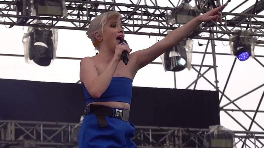 En un escenario iluminado, Carly Rae Jepsen canta con un micrófono, luciendo un conjunto azul y cabello rubio platino, mientras señala hacia arriba con su mano.