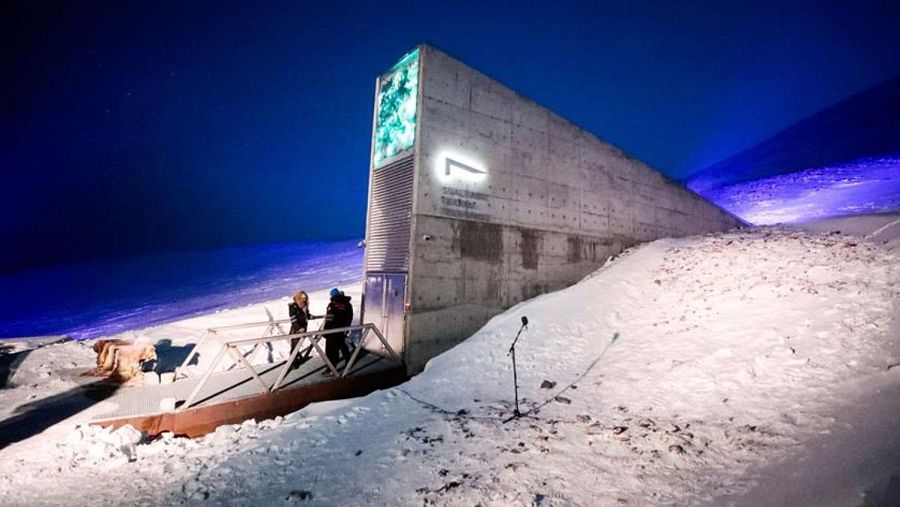 Un imponente edificio de hormigón, con una decoración artística y un logo luminoso, se alza bajo un cielo nocturno. Dos personas observan la estructura desde una pasarela metálica, en un entorno cubierto de nieve.