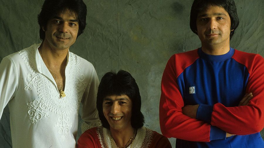 Fotografía de los tres integrantes de un grupo musical. Uno de ellos lleva una camisa blanca con bordados, otro una camisa roja y el tercero una sudadera azul con mangas rojas. El fondo es gris.