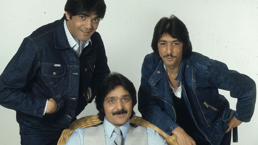 Tres hombres posan para una foto, uno sentado en una silla de mimbre, sobre un fondo blanco. Uno de ellos lleva una chaqueta vaquera azul, otro un chaleco gris y el tercero una chaqueta vaquera con cremallera.
