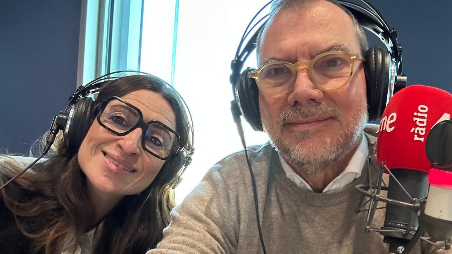 En un estudio de radio, dos personas sonríen a la cámara. Una mujer con gafas y cabello oscuro, y un hombre con gafas y cabello canoso, ambos con auriculares, se encuentran frente a un micrófono.