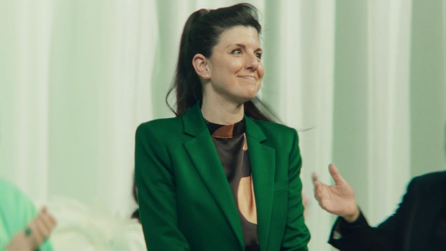 Alexina B, con el cabello recogido, posa con una chaqueta verde y una blusa estampada. El fondo es de color verde claro y se intuyen otras personas.