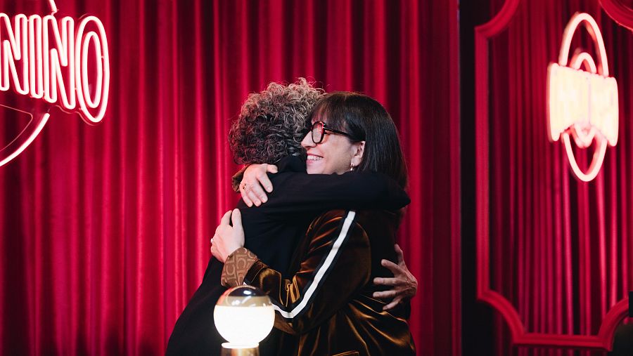 Judith Colell, cineasta, somriu mentre s'abraça amb una altra persona en un estudi amb cortines vermelles, portant ulleres i una jaqueta fosca amb una franja blanca. Al fons, un rètol de neó il·lumina l'espai.