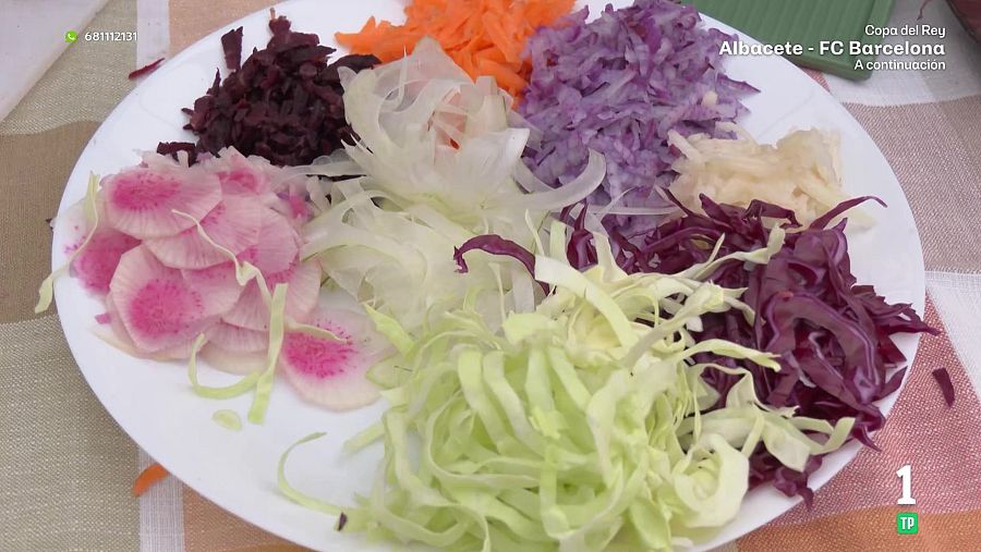 Ensalada con diversas verduras cortadas y dispuestas en un plato blanco, incluyendo rábanos, remolacha, zanahoria, cebolla, col lombarda, col blanca y col roja. La disposición es circular y colorida. Se observan elementos de texto y logos.