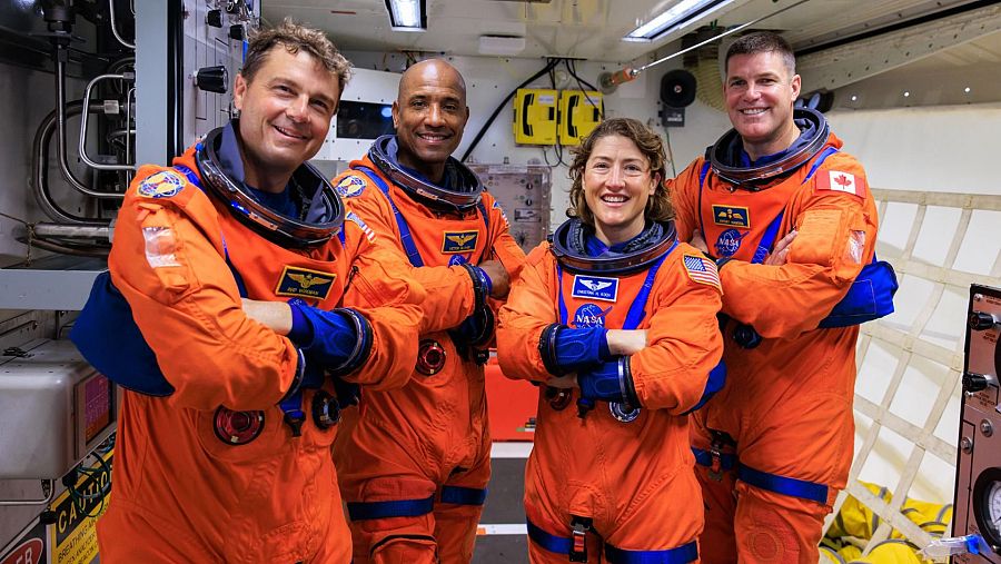 Cuatro astronautas con trajes espaciales naranjas y detalles azules posan sonriendo, posiblemente en un simulador de nave espacial. Dos de ellos llevan parches de banderas nacionales en sus trajes.