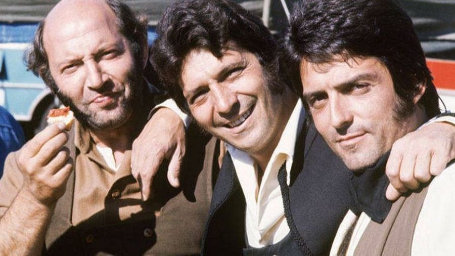 Tres hombres sonrientes posan juntos; uno come, otro sonríe ampliamente y el tercero tiene una expresión seria. Visten ropa similar y se encuentran frente a un fondo difuminado con un vehículo.