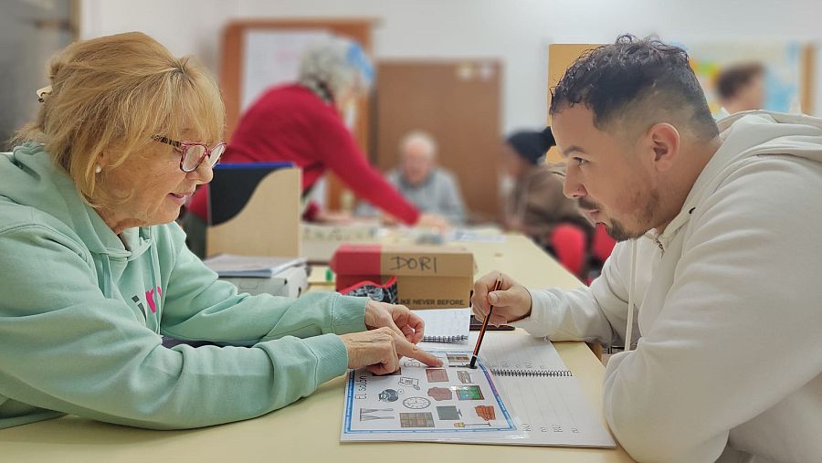 En una mesa, una voluntaria con gafas y sudadera verde del proyecto "¿Hablamos?" enseña vocabulario a un hombre joven con barba y sudadera blanca, utilizando una hoja con dibujos y un lápiz. Se observa una caja de cartón con la palabra "DORI".