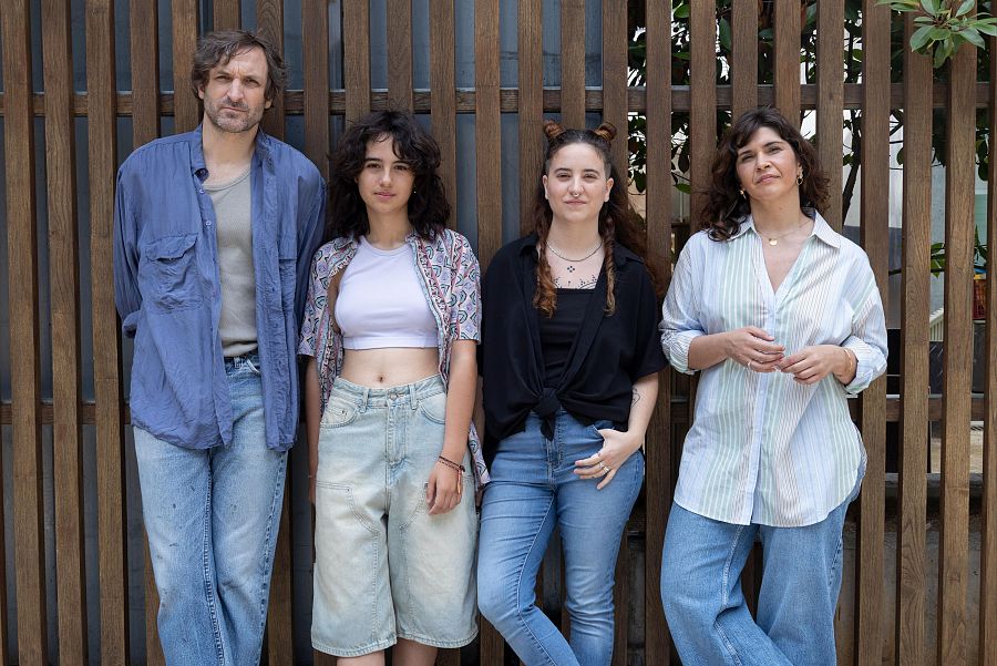 Cuatro personas posan frente a una pared de madera. Se distinguen un hombre con camisa azul, una joven con top blanco, otra con moños y camiseta negra, y una mujer con camisa de rayas, todos con jeans.
