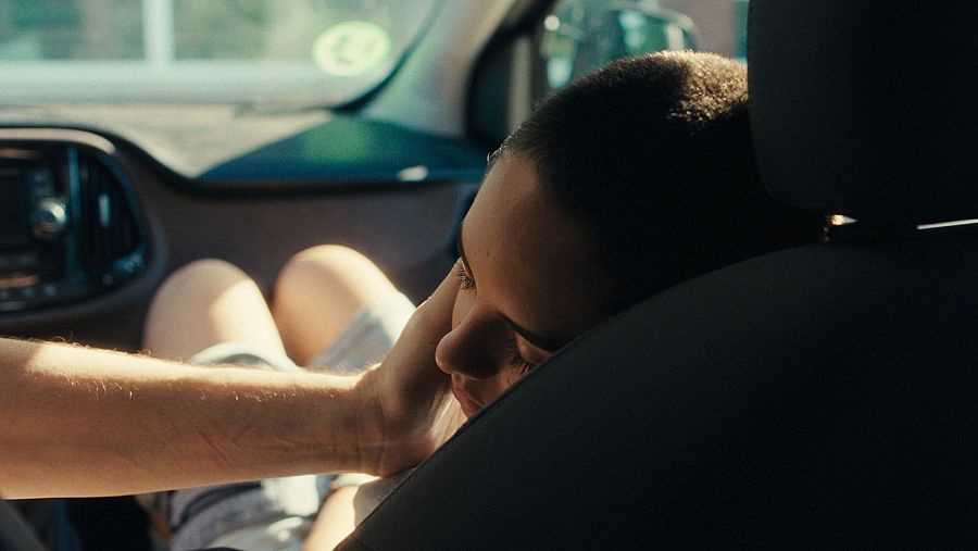 Primer plano de una persona con cabello oscuro recostada en el asiento trasero de un coche, con una mano tocando su rostro en un gesto de afecto. Se distingue parte del interior del vehículo.