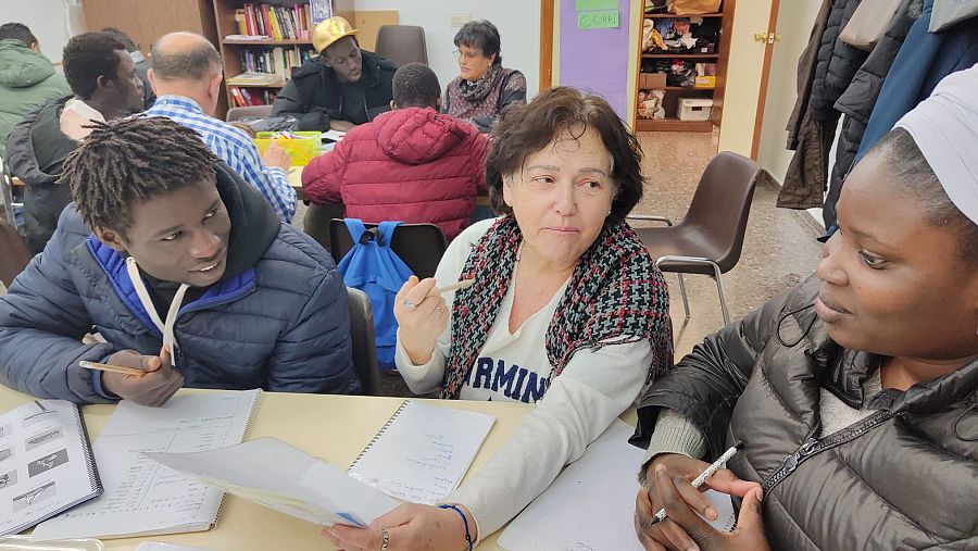 En un aula, dos estudiantes, uno con chaqueta azul y otro con rastas, escriben. Una mujer con un pañuelo y una camiseta con la palabra "ARMIN" explica algo, mientras otros observan. Se ven estanterías con libros.