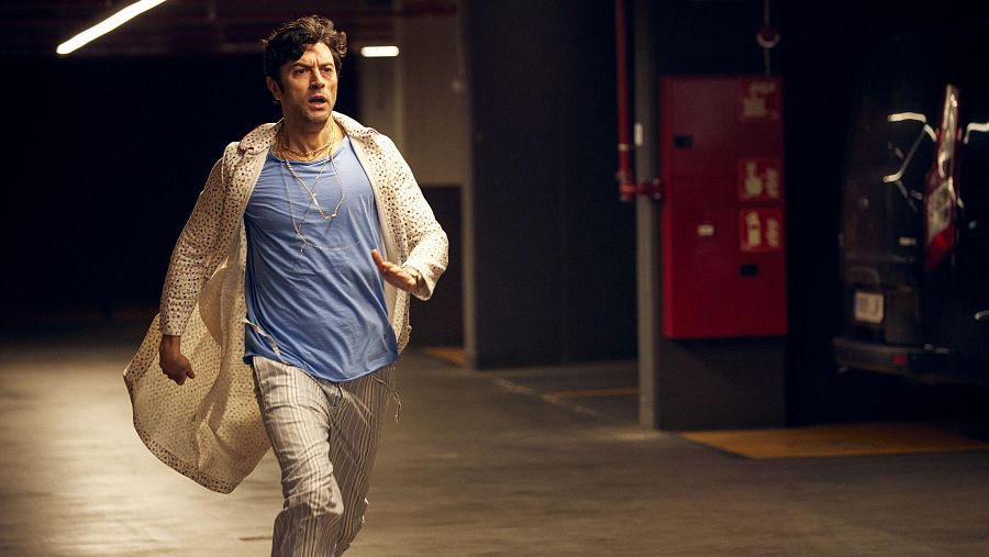 Un hombre con cabello rizado y oscuro, vestido con camiseta azul, pantalones a rayas, cárdigan crema y collares dorados, corre con expresión de sorpresa en un aparcamiento subterráneo, con una furgoneta y una caja de incendios al fondo.