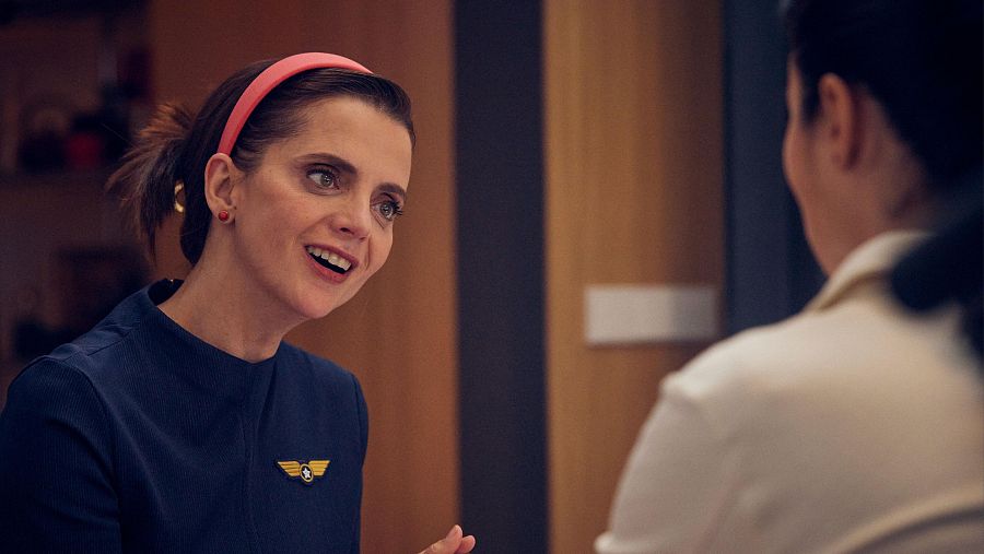 Macarena Gómez, con una diadema rosa y un uniforme azul oscuro, sonríe y gesticula mientras conversa con alguien que está de espaldas, en un ambiente con fondo de madera.