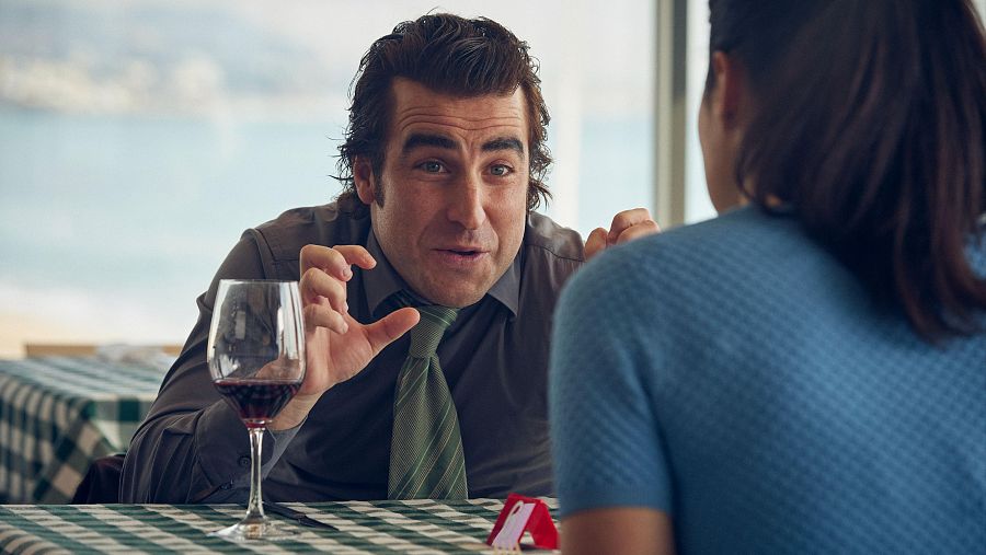 Marco Cáceres, interpretando a Lorenzo, conversa con otra persona en una escena de 'El fantasma de mi mujer'. Viste camisa gris y corbata verde, mientras gesticula con las manos. En la mesa hay una copa de vino tinto.