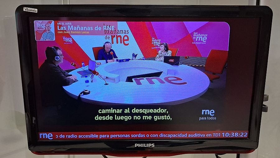 Una emisión en directo del programa "Las Mañanas de RNE" se muestra en una pantalla de televisión. En la imagen, se distingue una mesa con el logo de la emisora y tres personas, además de subtítulos informativos.