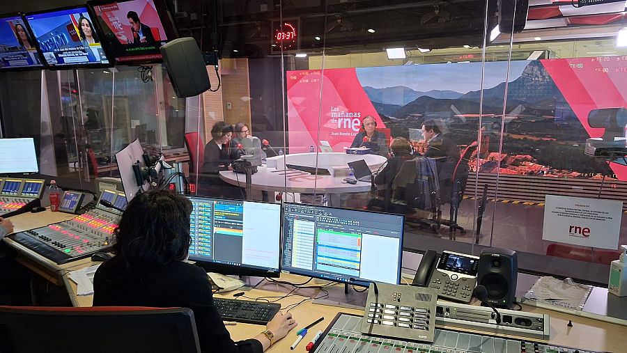 En una sala de control de radio, una persona trabaja con equipos técnicos, como mesas de mezclas y monitores, mientras se transmite una entrevista en el estudio contiguo. Se observa un reloj digital marcando la hora.