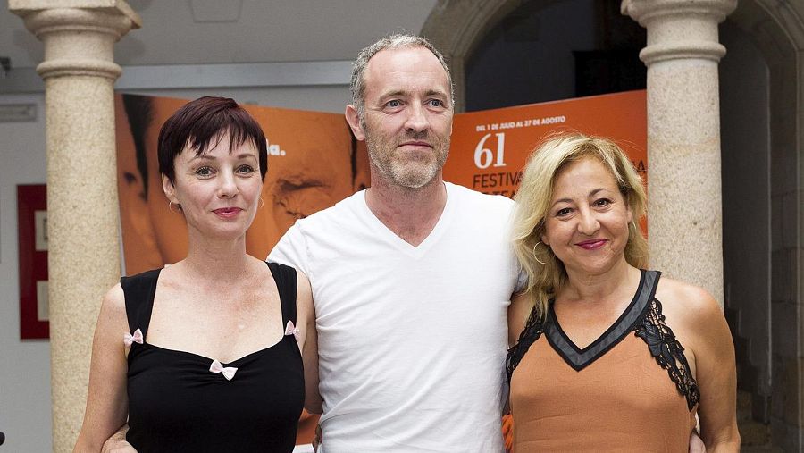 Tres personas posan frente a un cartel naranja. Dos mujeres y un hombre. Una de las mujeres lleva una blusa negra con lazos rosas y la otra una blusa ocre con encaje negro. El cartel anuncia un festival de teatro.