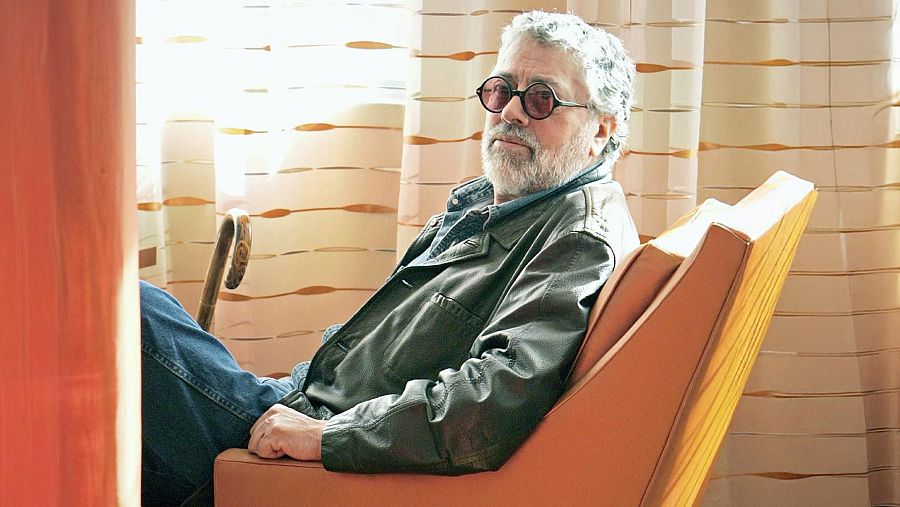 Facundo Cabral, con gafas oscuras y vestimenta casual, posa sentado junto a un bastón. El fondo presenta una cortina con diseño horizontal.