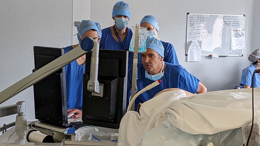 Un equipo médico, con gorros y mascarillas, se concentra en monitores que muestran imágenes de un procedimiento. Un paciente cubierto y un brazo robótico son visibles en primer plano, mientras que una pizarra con anotaciones se encuentra al fondo.