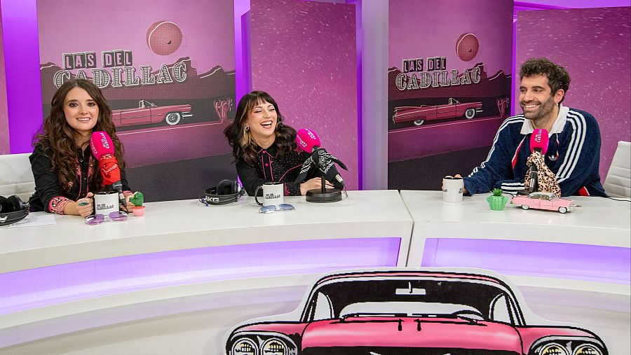 Tres mujeres, posiblemente locutoras, están sentadas frente a micrófonos en un estudio con decoración de color rosa y elementos relacionados con un Cadillac. En la mesa hay tazas, auriculares y un cactus.