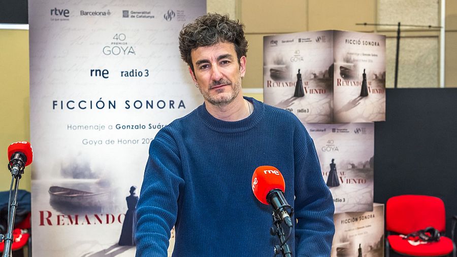 Víctor Clavijo, con barba y jersey azul, se encuentra frente a carteles promocionales que anuncian "FICCIÓN SONORA" y "REMANDO AL VIENTO", con logos de radio y premios. Hay micrófonos rojos y una silla en el fondo.