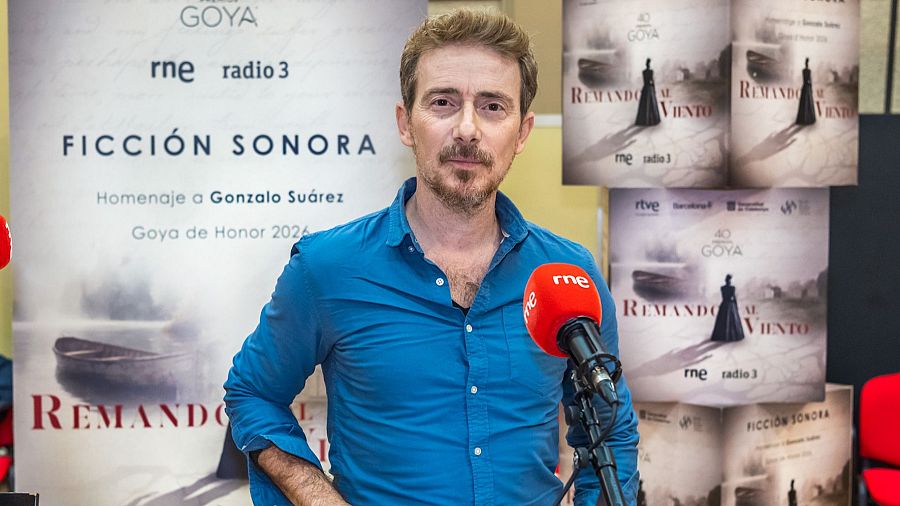Víctor Clavijo, con camisa azul, se encuentra frente a un micrófono rojo, con carteles promocionales de "Remando al Viento" detrás, incluyendo logos de premios y emisoras.