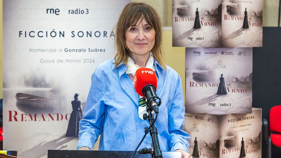 Natalie Poza, con camisa azul y pañuelo, posa frente a un atril con micrófono, probablemente durante los ensayos de una producción radiofónica. Se observan carteles promocionales con una imagen de una mujer y logos de RTVE.