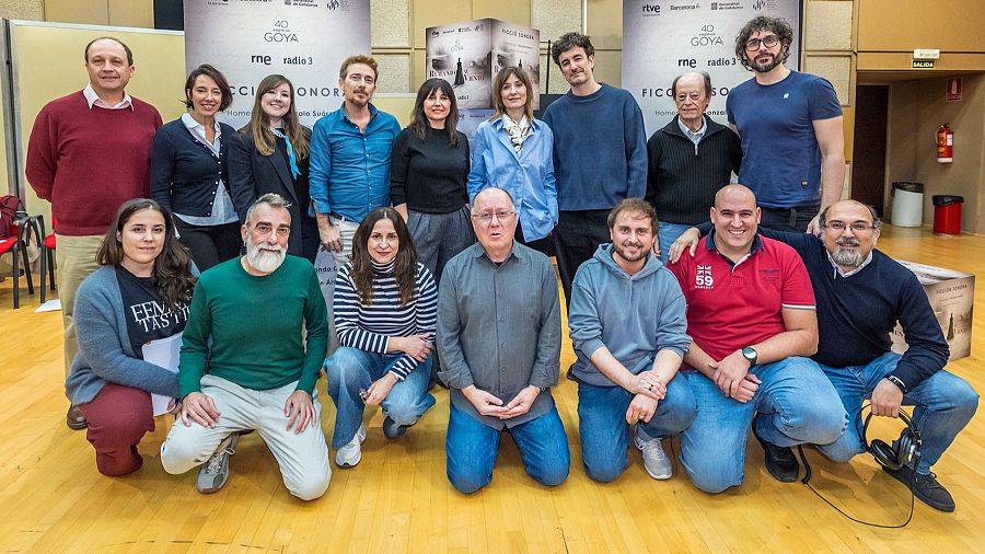 Un grupo de dieciséis personas posan para la foto. Se observan hombres y mujeres con diversas prendas, como camisas, jerseys y chaquetas. En el fondo, se distinguen carteles y la portada de un libro.