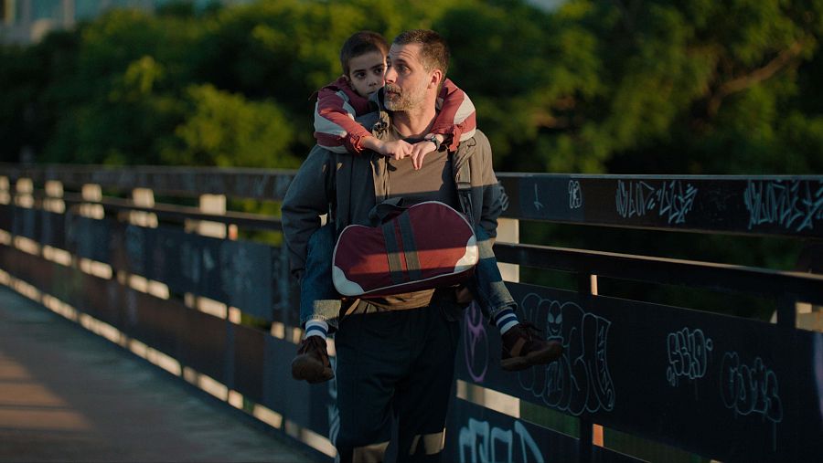 En un puente con barandillas cubiertas de grafitis, un hombre con barba lleva a un niño pequeño sobre sus hombros. El hombre viste ropa deportiva y el niño una chaqueta a rayas.