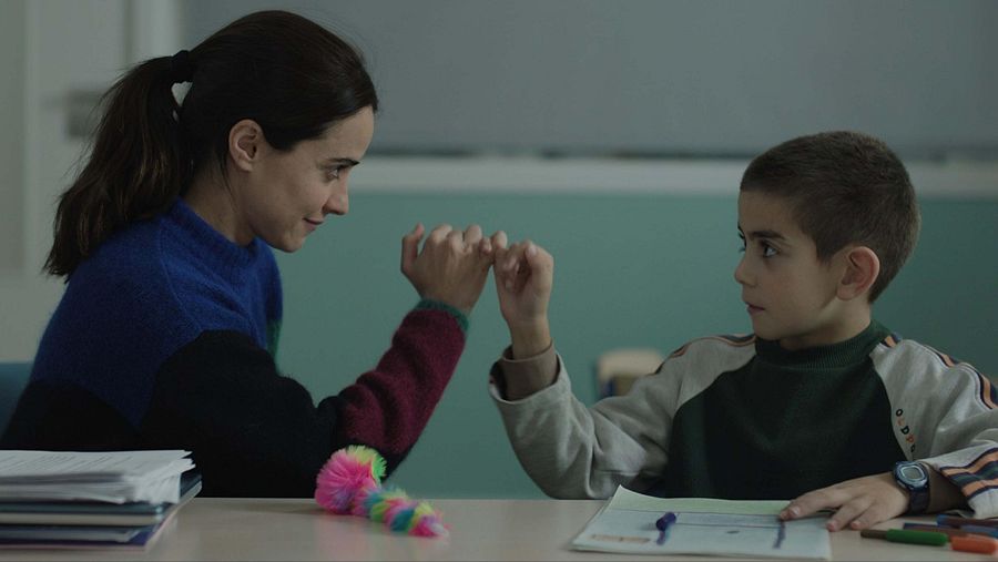 Un fotograma de la película muestra a una mujer y un niño sentados a una mesa. La mujer, con el pelo recogido, entrelaza su dedo meñique con el del niño, que lleva una camiseta de manga larga. En la mesa hay libros, un dibujo y un juguete.