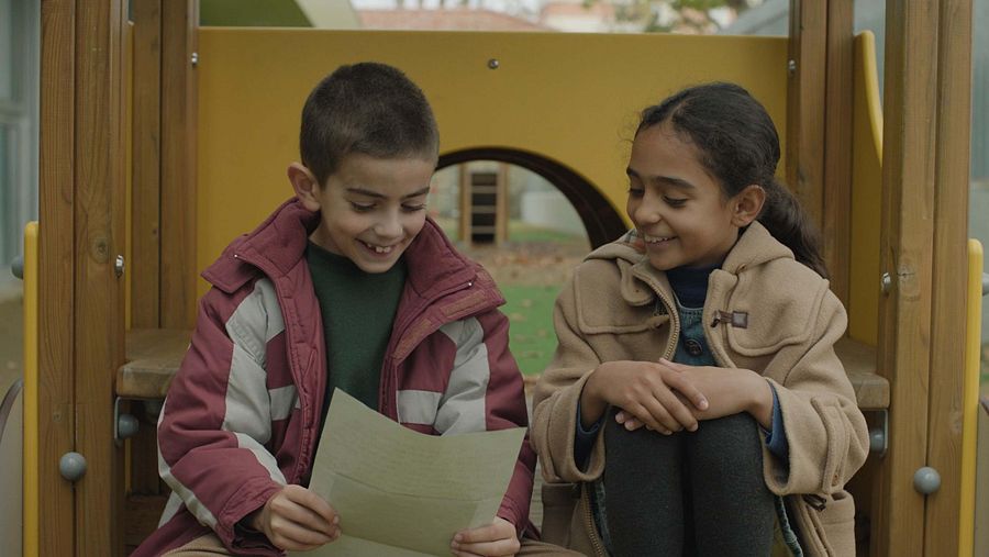 En un parque infantil, un niño con chaqueta roja y una niña con abrigo color camel interactúan. Ambos sonríen, con el niño mirando un papel y la niña observándolo a él. El entorno es al aire libre, con una estructura de juegos de madera y metal.