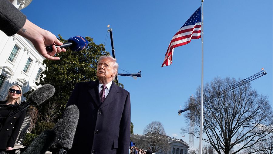 En una escena al aire libre, Donald Trump, con vestimenta formal, habla ante micrófonos. Una mujer con gafas de sol está presente, y al fondo se observa un edificio blanco con la bandera de Estados Unidos.