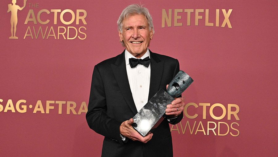 Harrison Ford, vestido con un esmoquin negro, sonríe sosteniendo un premio plateado con forma de máscara teatral, en un evento donde se aprecia el logo de "The Actor Awards" y "SAG-AFTRA".