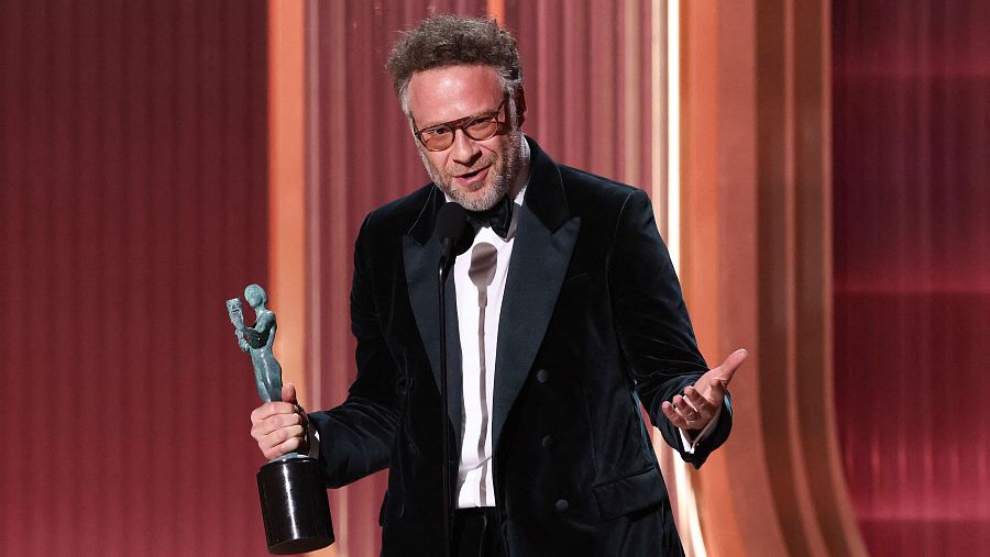 Seth Rogen, con gafas y un traje de terciopelo negro con pajarita, sostiene un premio en una ceremonia con fondo rojizo y líneas verticales.