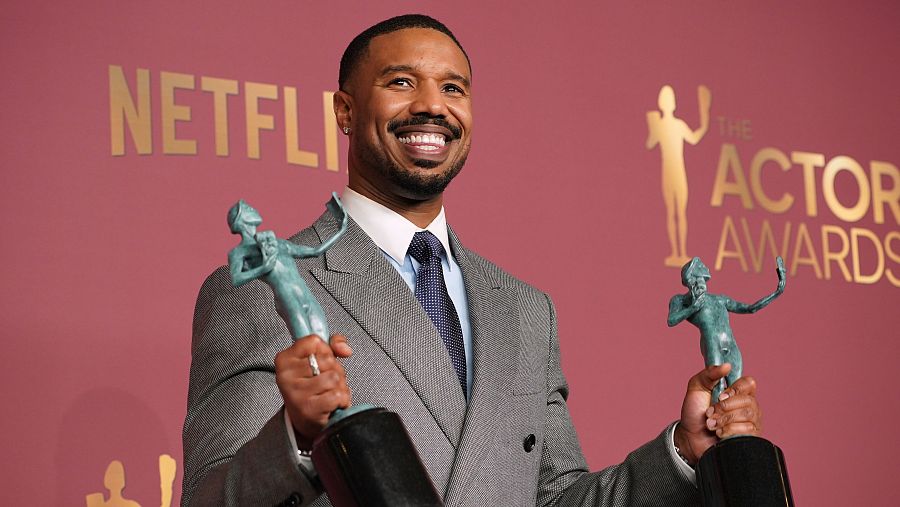 Michael B. Jordan, con un traje gris y corbata azul, posa sonriente con dos estatuillas doradas frente a un fondo con logos. El actor lleva un anillo plateado en su mano izquierda.