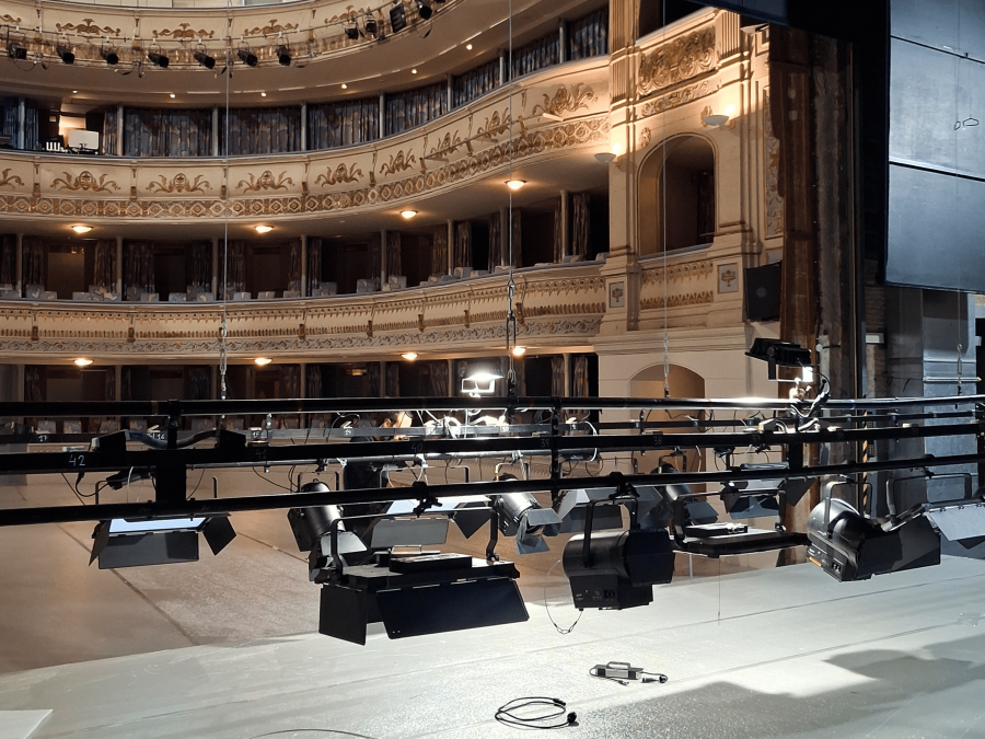 Interior de un teatro con balcones decorados y luces cálidas. El escenario, equipado con luces colgantes y focos, muestra cables y conectores en el suelo, sugiriendo preparativos.