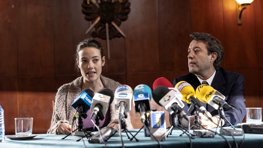 Dos personas, una mujer con cárdigan y un hombre con traje, están sentadas frente a una mesa con micrófonos, posiblemente en una rueda de prensa. Se observa una botella de agua, un vaso y una grabadora sobre la mesa.