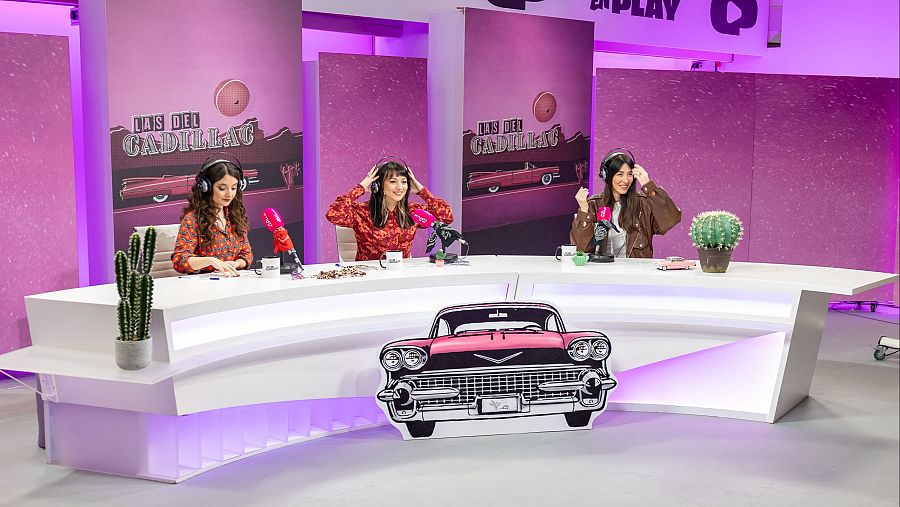 Tres mujeres, María Canet, Raquel Elices y Natalia Ferviú, están sentadas frente a micrófonos en una mesa con iluminación morada, grabando un podcast. Detrás, paneles con el logo de un programa y un coche rosa.