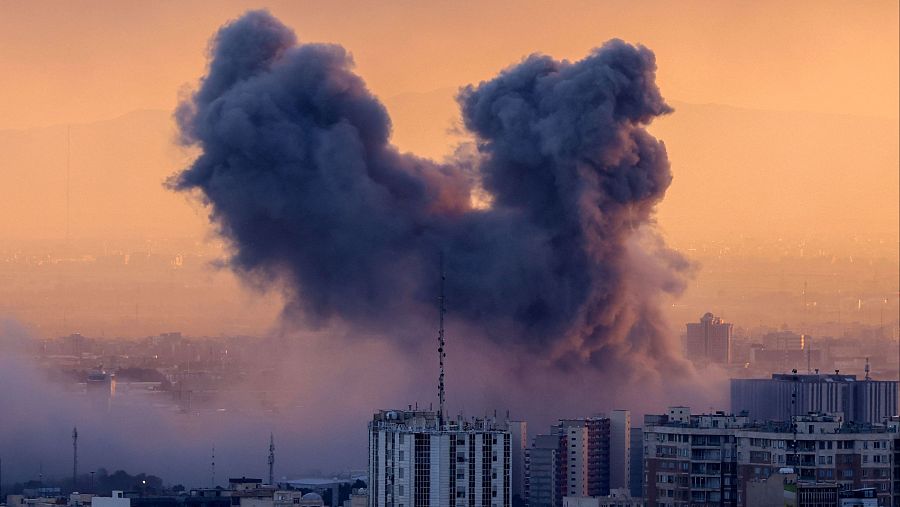 Una gran explosión en una zona urbana crea una densa columna de humo sobre edificios altos, con una ciudad y montañas visibles al fondo, bajo un cielo anaranjado.