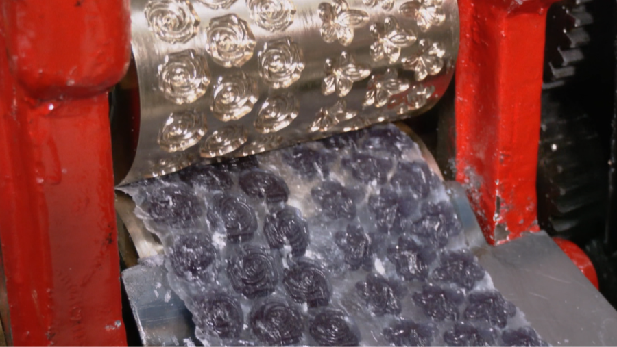 Una máquina industrial de metal rojo utiliza un molde con forma de flor para dar forma a caramelos de color violeta translúcido. Los caramelos ya formados se acumulan en la parte inferior, listos para su separación.