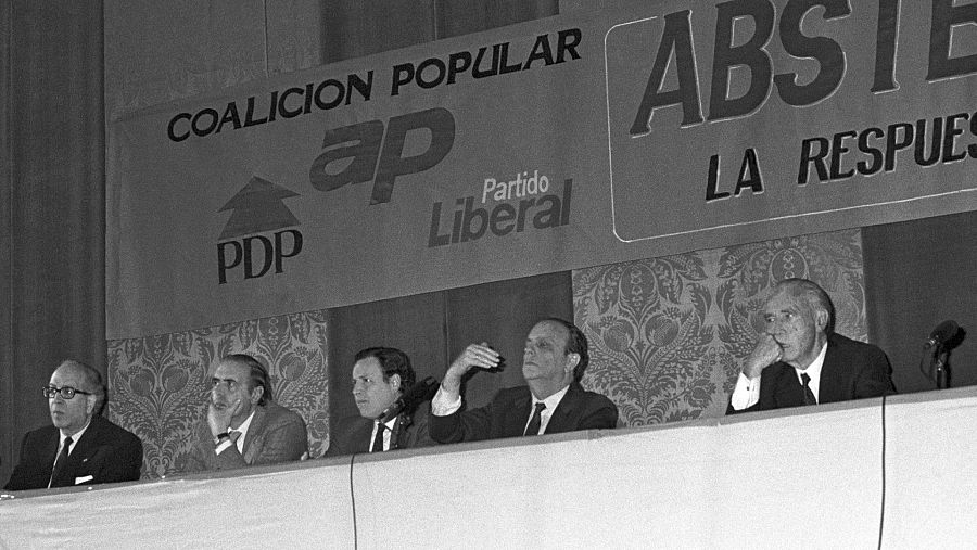 Una fotografía en blanco y negro muestra a cinco hombres sentados tras una mesa, con un cartel político al fondo. Uno de ellos es Manuel Fraga, presidente de una formación política.