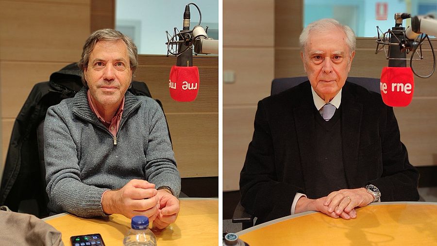 Dos hombres, uno con jersey gris y otro con traje negro, están sentados frente a micrófonos en un estudio de grabación. En la mesa, hay un teléfono móvil y una botella de agua. El fondo es una pared con paneles de madera.