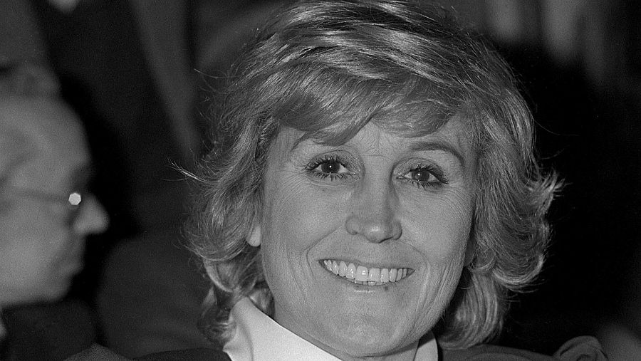 En la fotografía en blanco y negro, se aprecia un primer plano de una mujer con cabello corto y ondulado, sonriendo. Se distingue la presencia parcial de otra persona con gafas en el margen izquierdo.