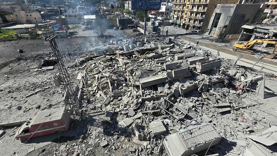 Una escena de destrucción revela escombros de edificios, con fragmentos de estructuras y un vehículo dañado. Una excavadora amarilla trabaja entre los restos, mientras que al fondo se ven edificios parcialmente destruidos.