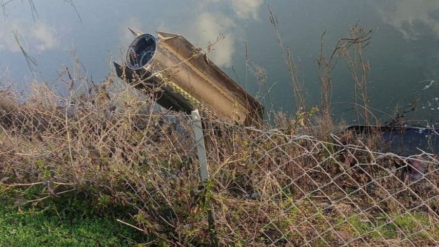 Restos de un misil dañado yacen en el agua cerca de la orilla, con vegetación seca y una valla metálica en primer plano. La imagen sugiere un ataque reciente en una zona costera.