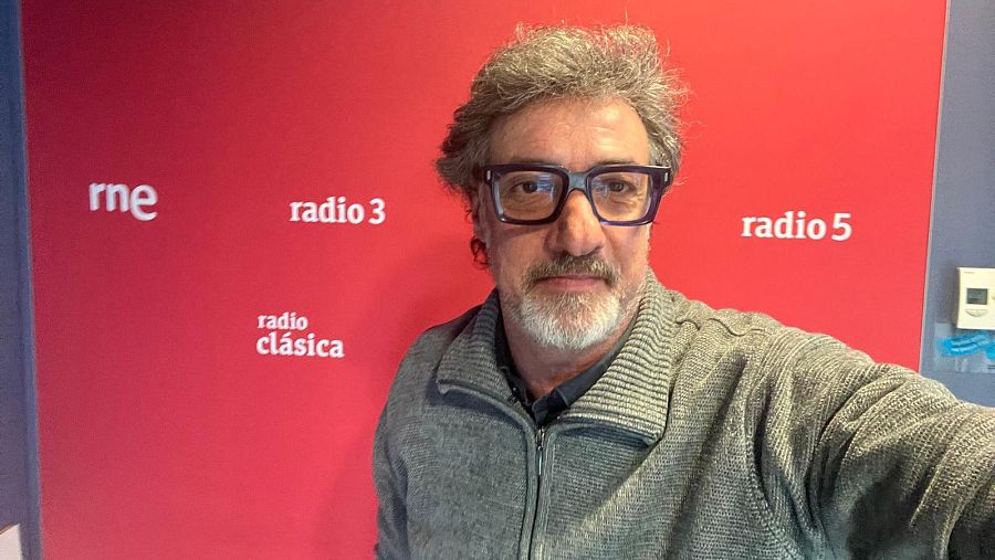 Gerard Jofra, con gafas y chaqueta de punto, posa frente a una pared con logotipos de emisoras de radio, posiblemente en un estudio.