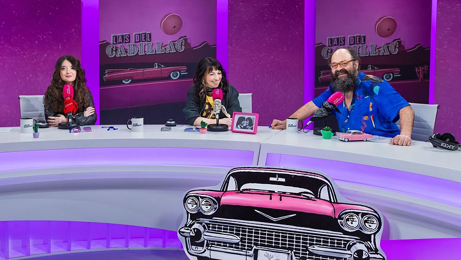 Tres personas, María Canet, Raquel Elices e Ignatius Farray, están sentadas en una mesa frente a micrófonos rojos, con carteles que anuncian "LAS DEL CADILLAC" detrás. En la mesa se observan tazas, un cactus y un coche de juguete.