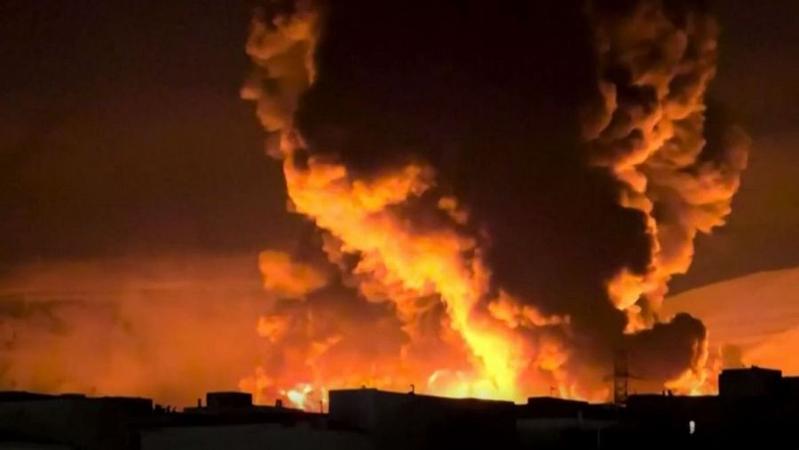 Un incendio de gran envergadura consume una zona, generando una columna de humo oscuro y llamas que se elevan hacia el cielo. La imagen muestra edificios en la parte inferior, indicando un origen urbano o industrial del fuego.