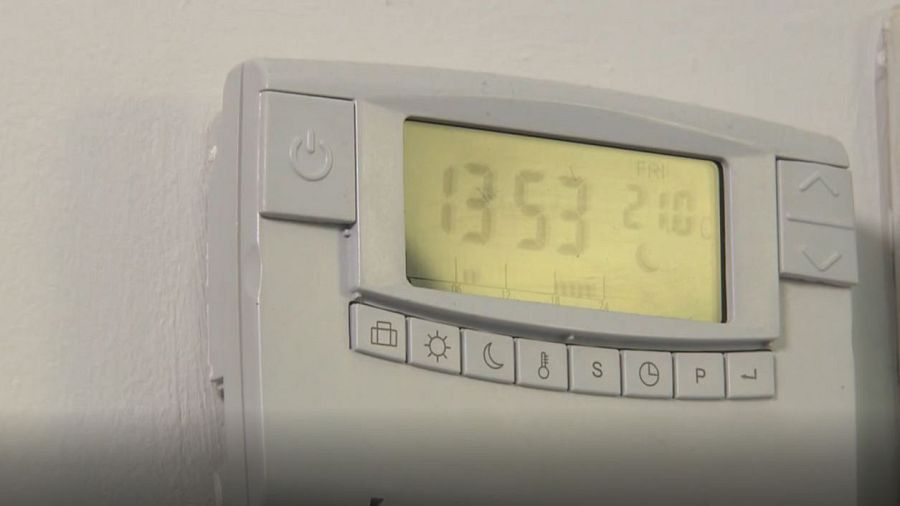 Un termostato digital blanco, con pantalla LCD, muestra la hora (13:53), el día (viernes) y la temperatura (21 grados Celsius). Cuenta con botones con símbolos para controlar sus funciones.